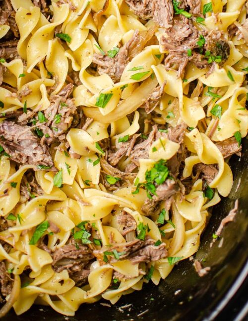 Closeup of pulled beef in the slow cooker with egg noodles with parsley sprinkled over top.