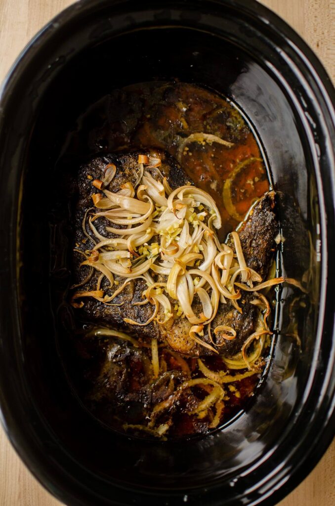 Cooked beef in the slow cooker insert, with caramelized onions on top.