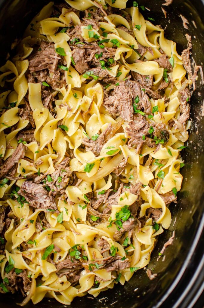 Pulled beef and noodles in a slow cooker insert with parsley sprinkled on top.