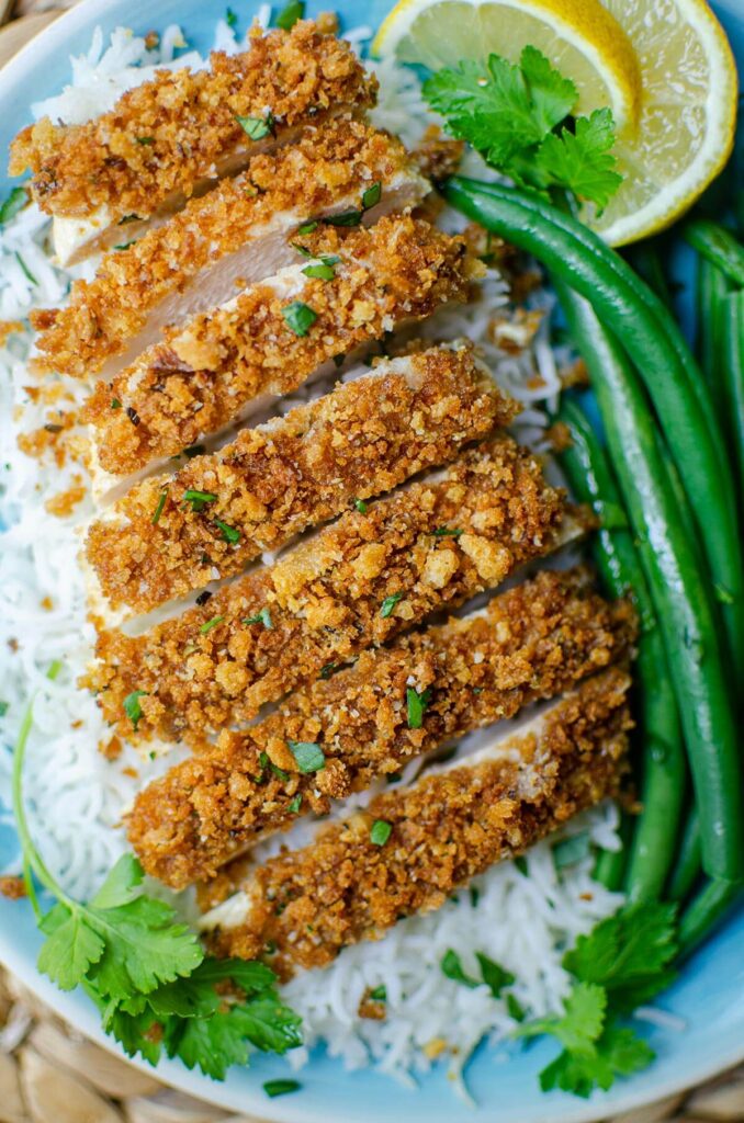 Sliced panko chicken on a bed of rice with green beans. Garnished with lemon and parsley.