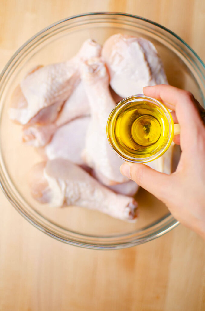Olive oil being poured over chicken drumsticks in a glass bowl.