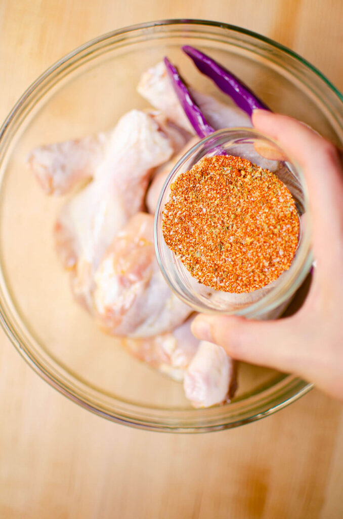 Spice rub in a glass bowl being poured over chicken drumsticks in a larger glass bowl.