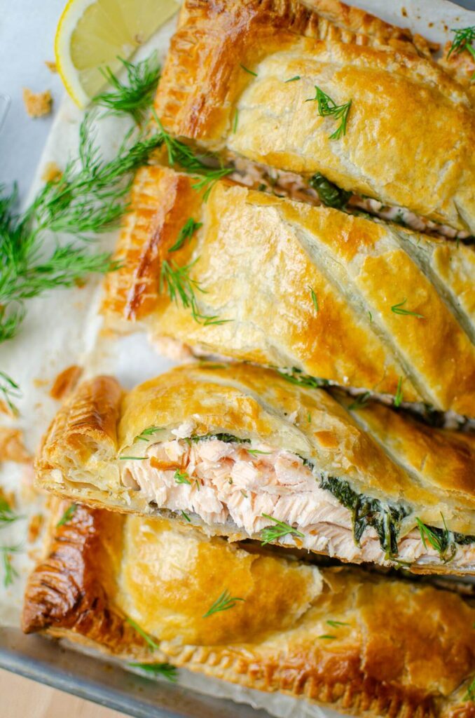 Closeup on the top half of four pieces of salmon wellington on a baking sheet with dill and lemon.