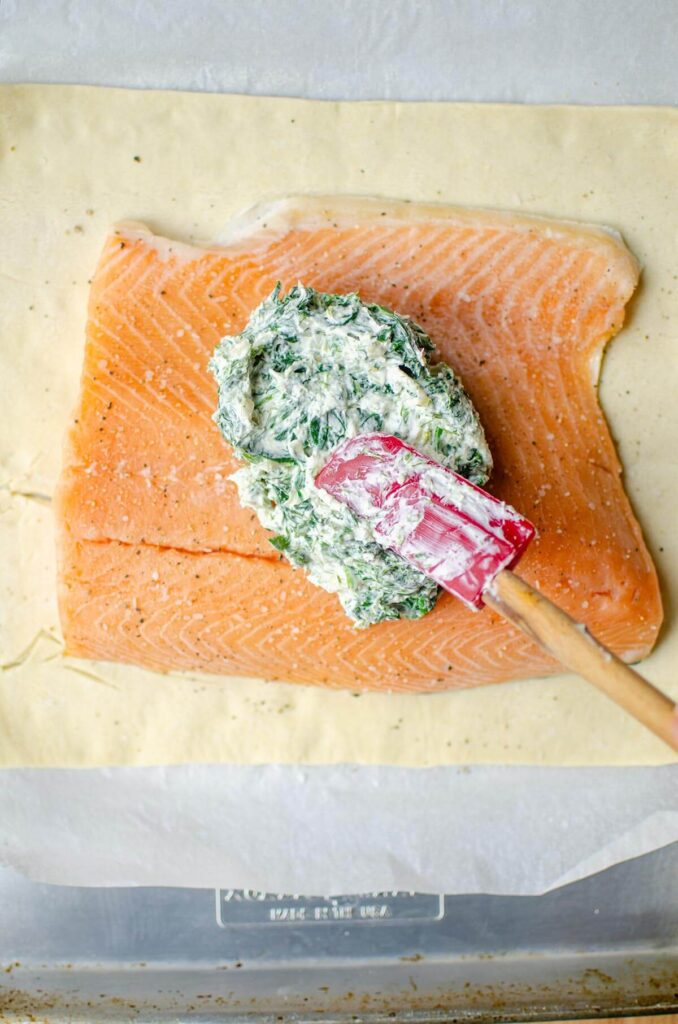A pile of spinach filling on the center of the peice of salmon with a spatula spreading it around.