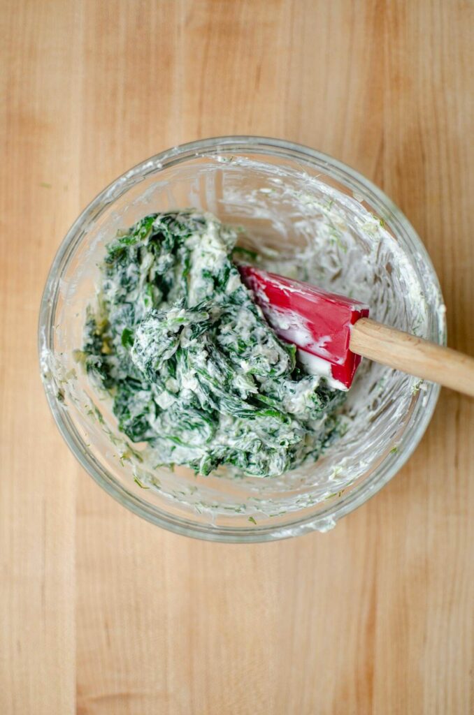 Spinach filling in a glass bowl being mixed with a red spatula.