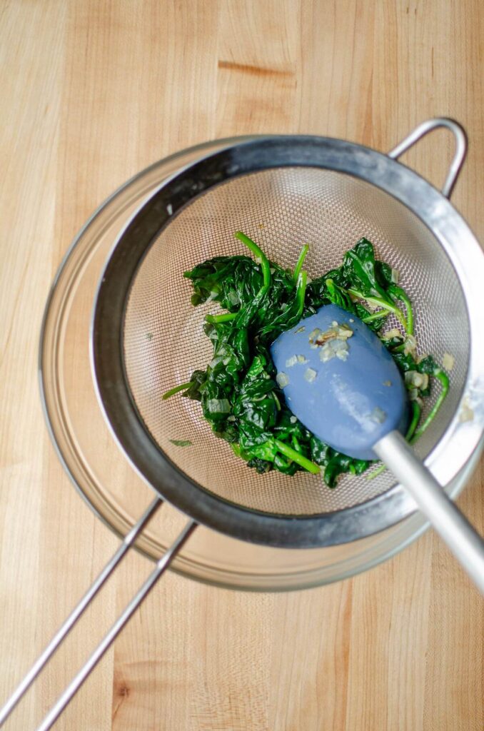 Cooked spinach in a sieve over a glass bowl, with a blue spatula pressing down on the spinach mixture, releasing the liquid.