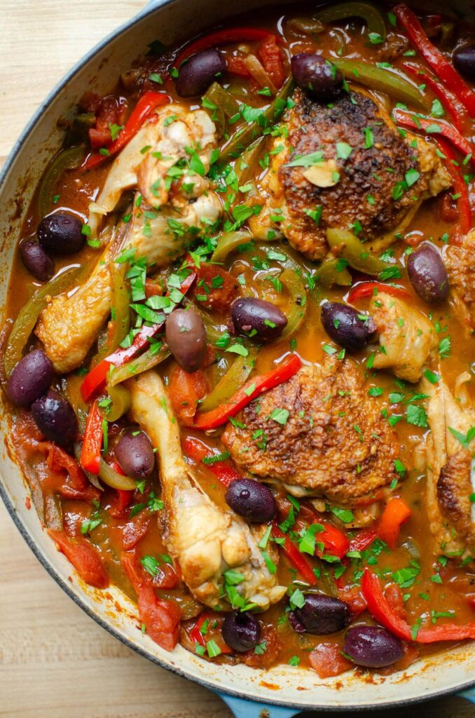 Half a braising dish with cooked chicken, peppers, and onions with olives and parsley on top.