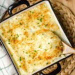 Closeup of twice baked mashed potatoes in a baking dish with a wooden spoon taking a serving.