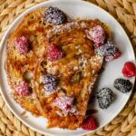 Two slices of french toast on a white plate with berries and maple syrup.