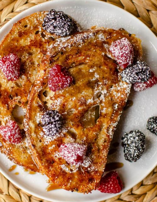 Two slices of french toast on a white plate with berries and maple syrup.