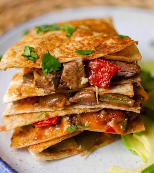 Stack of quesadillas on top of each other with sliced avocado on a plate.